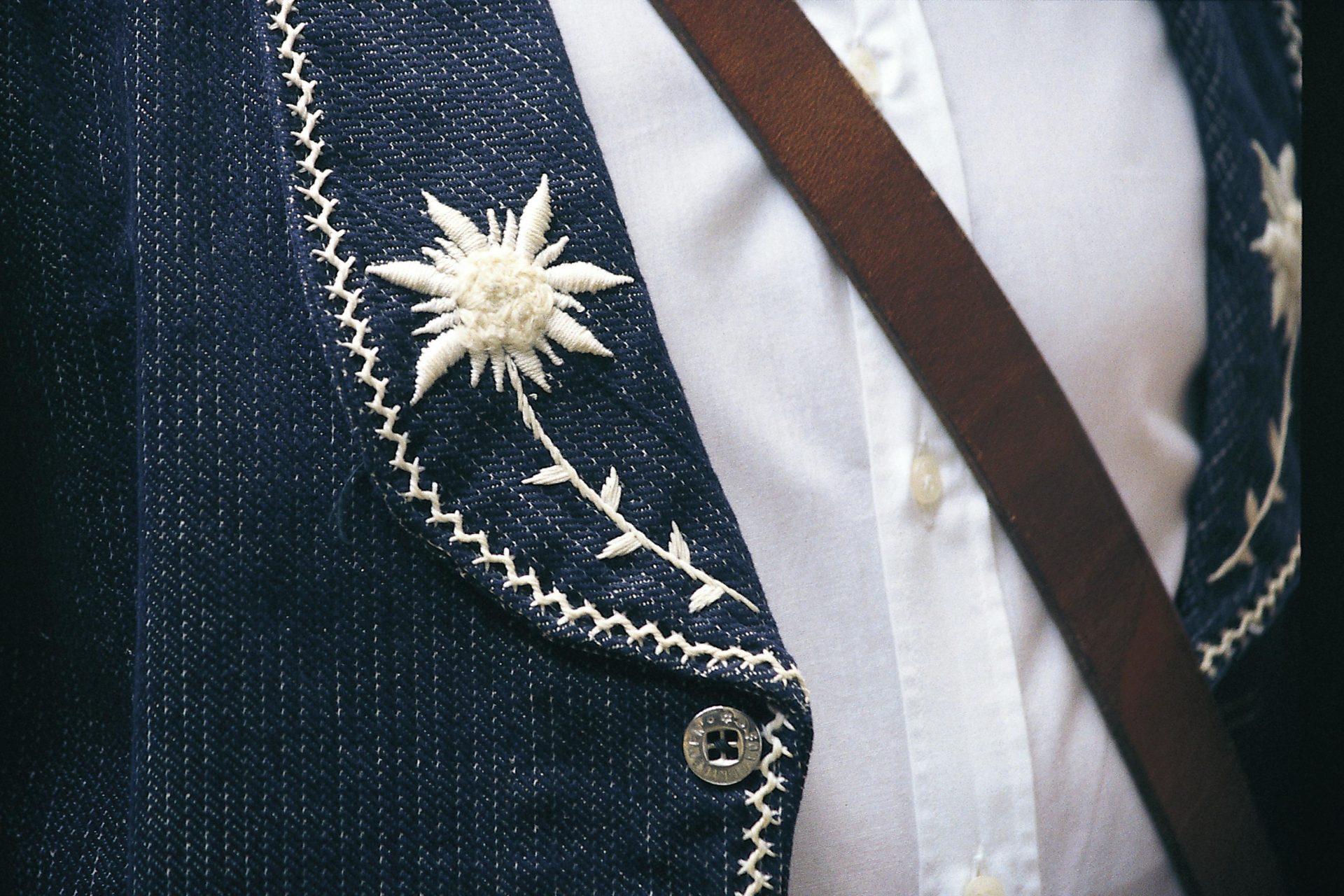 Detailansicht einer blauen Jacke mit aufwendiger weißer Blumenstickerei und Lederriemen, die ein elegantes Modedesign präsentiert.